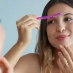 Woman using a pink facial razor to dermaplane her forehead, smiling softly, showcasing Dermaplaning in Missoula, MT.