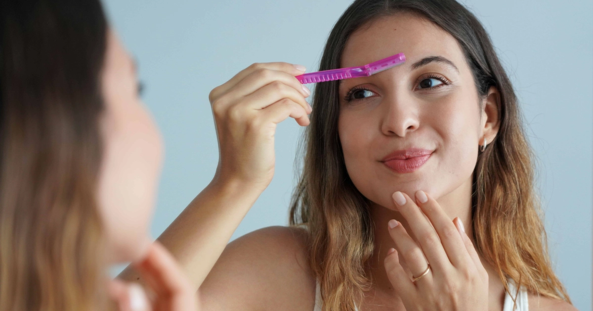 Woman using a pink facial razor to dermaplane her forehead, smiling softly, showcasing Dermaplaning in Missoula, MT.